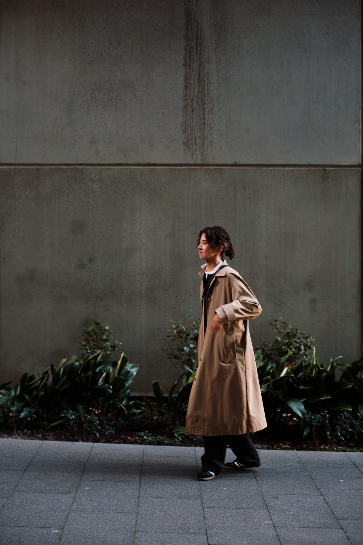 Julia walking past a concrete wall in a trench coat